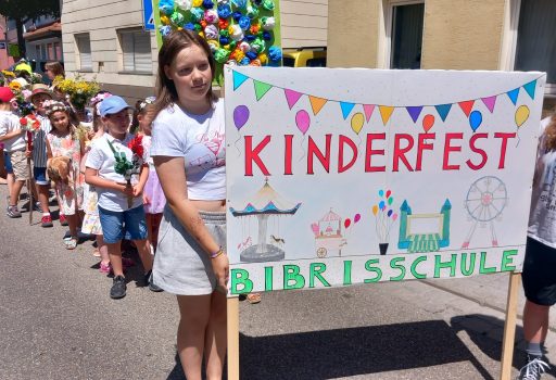 Eine Gruppe von Kindern steht draußen in einer Reihe und hält ein buntes Schild mit der Aufschrift „Kinderfest Bibrisschule“ hoch, das mit Zeichnungen von Luftballons, einem Riesenrad und Karnevalsszenen verziert ist. Die Sonne scheint strahlend.