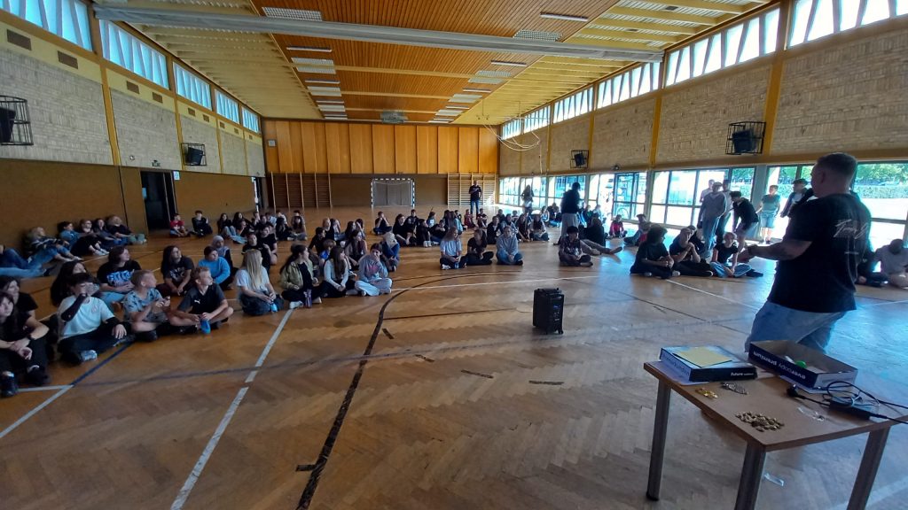 Eine Gruppe von Menschen sitzt auf dem Boden einer Turnhalle und blickt zu einem Redner vorne im Raum, der neben einem Tisch mit elektronischen Geräten steht. An den Wänden befinden sich große Fenster, durch die Tageslicht hereinfällt.