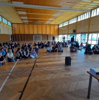 Eine Gruppe von Menschen sitzt auf dem Boden einer Turnhalle und blickt zu einem Redner vorne im Raum, der neben einem Tisch mit elektronischen Geräten steht. An den Wänden befinden sich große Fenster, durch die Tageslicht hereinfällt.