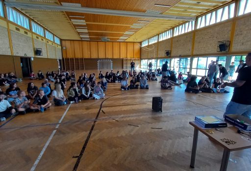 Eine Gruppe von Menschen sitzt auf dem Boden einer Turnhalle und blickt zu einem Redner vorne im Raum, der neben einem Tisch mit elektronischen Geräten steht. An den Wänden befinden sich große Fenster, durch die Tageslicht hereinfällt.
